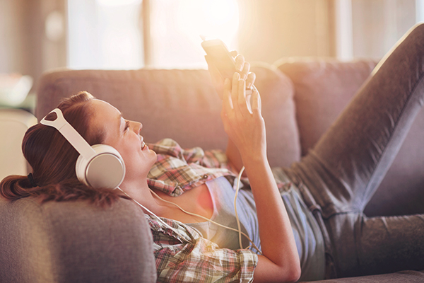 Im Wohnzimmer eine Frau trägt Kopfhörer, liegt auf dem Couch und schaut auf einem Smartphone