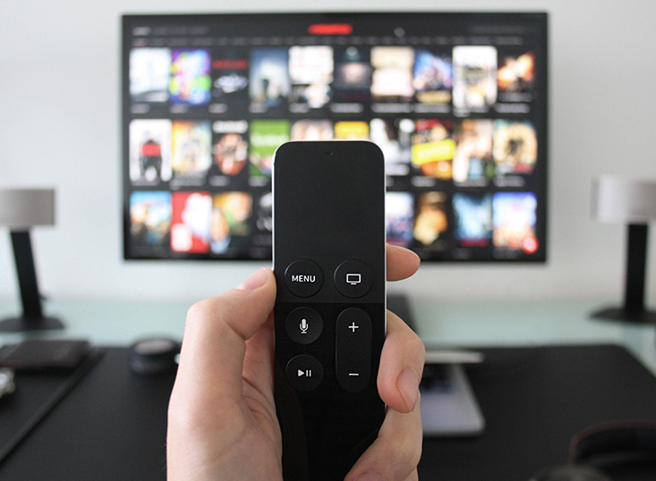 Eine Hand hält die Fernbedienung eines Streaming-Geräts in die Höhe. Im Hintergrund ist ein Smart TV mit der Benutzeroberfläche eines Streaming-Dienstes zu erkennen.