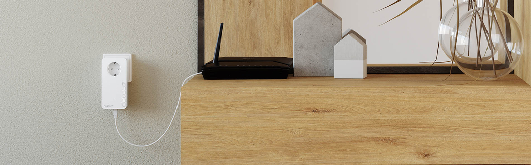 Kleiner Schrank aus Holz im Wohnzimmer. Drauf ist ein schwarzer WLAN Router und andere Wohnzimmer-Dekos. Neben dem Schrank ist ein devolo Magic LAN Powerline-Adapter in einer Steckdose an der Wand angeschlossen. Der devolo Magic Adapter ist mit einem Kabel an dem Router angeschlossen.