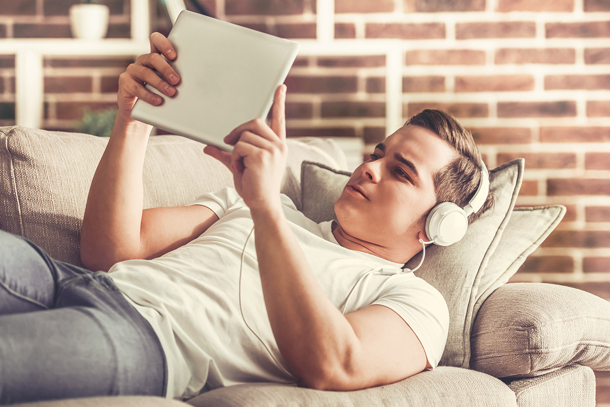 Junge liegt auf dem Couch. Er trägt Kopfhörer und schaut fokussiert auf dem Tablet.