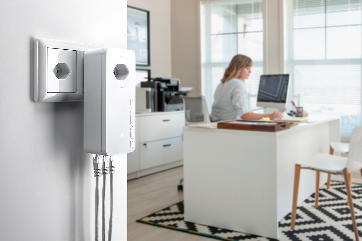 Frau im Büro im Hintergrund. An der Wand in einer Steckdose ist ein devolo dLAN 1200 triple angeschlossen. Mit dem Powerline Adapter sind 3 LAN Kabeln verbunden.