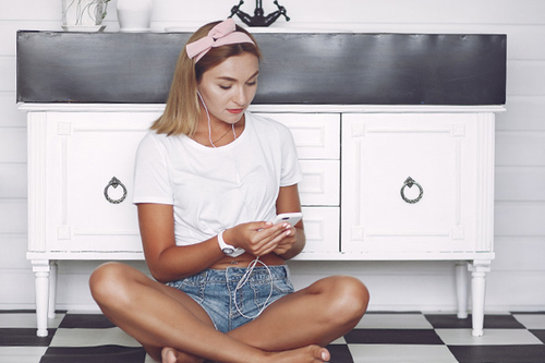 Frau badet in der Badewanne. Sie trägt Kopfhörer und hat ein Getränk in einer Hand und ein Smartphone in die andere Hand.