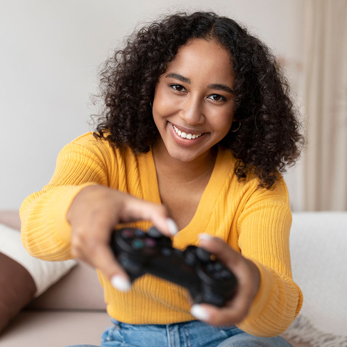 Eine Frau sitzt auf der Couch und reckt lächelnd den Gaming-Controller in die Kamera.