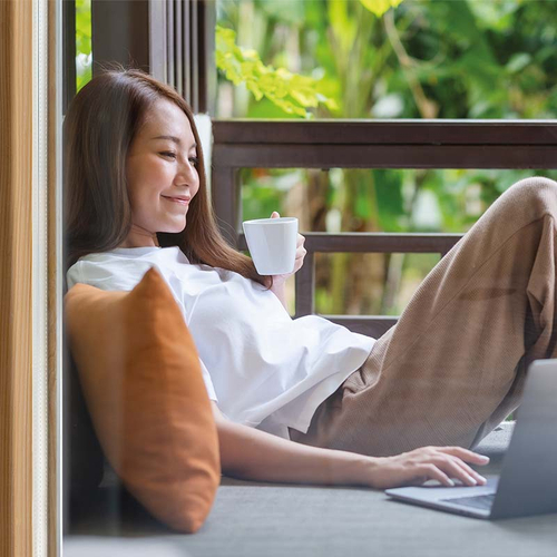 Eine Frau macht es sich mit Tasse und Laptop auf dem Balkon gemütlich und surft im Internet.