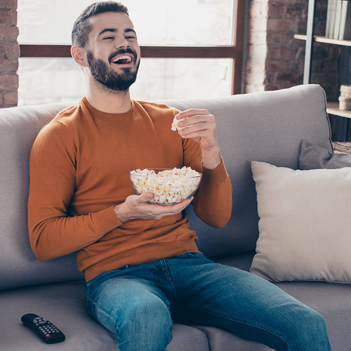 Ein Mann sitzt mit Popcorn und Fernbedienung auf der Couch und genießt lachend das Streaming-Angebot.