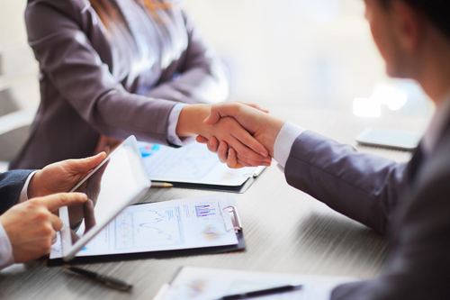 A meeting table with lots of documents on it and two people shaking hands. Another person is operating a tablet.