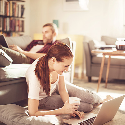 Mann und Frau sitzen jeweils auf dem Couch und Boden und streamen auf dem Tablet und Laptop.