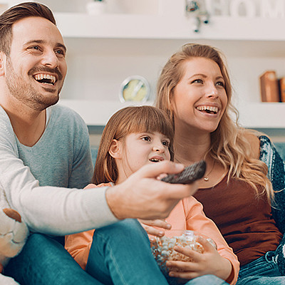 Familie sitzt auf dem Couch im Wohnzimmer und schaut Fern. Der Mann hält die Fernbedienung.